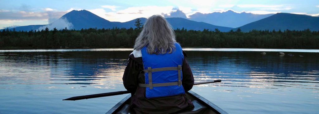 Back of a woman at the front of a canoe on a still lake, facing forested shoreline and distant mountains